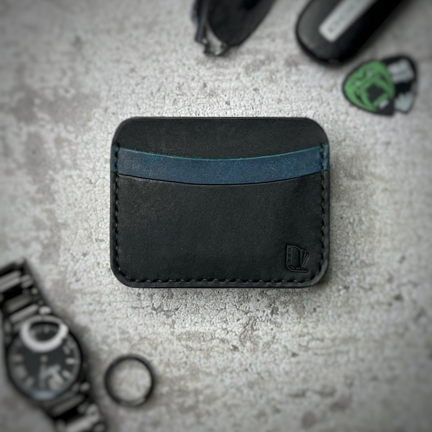 A leather cardholder with a black body and a blue strip across the top, featuring a minimalist design. The cardholder is placed on a gray textured surface, surrounded by a black watch, a black ring, and some keys and other small objects in the background.