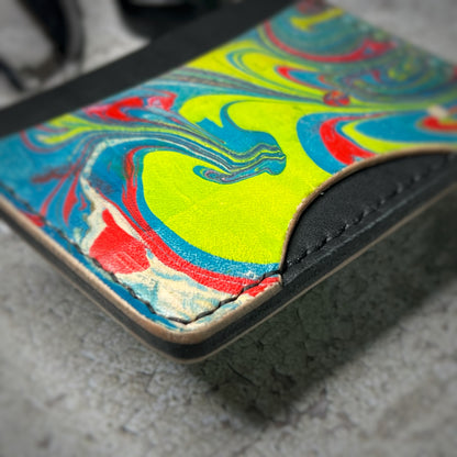 Close-up view of a colorful leather cardholder with a black upper section and a vibrant, swirling pattern in yellow, blue, red, and green on the lower section. The cardholder is placed on a gray textured surface, highlighting the stitching details.