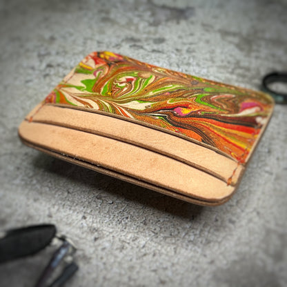 A leather cardholder with a natural tan upper section and a colorful, swirling pattern in green, red, and brown on the lower section, featuring a slightly angled view. The cardholder is placed on a gray textured surface.