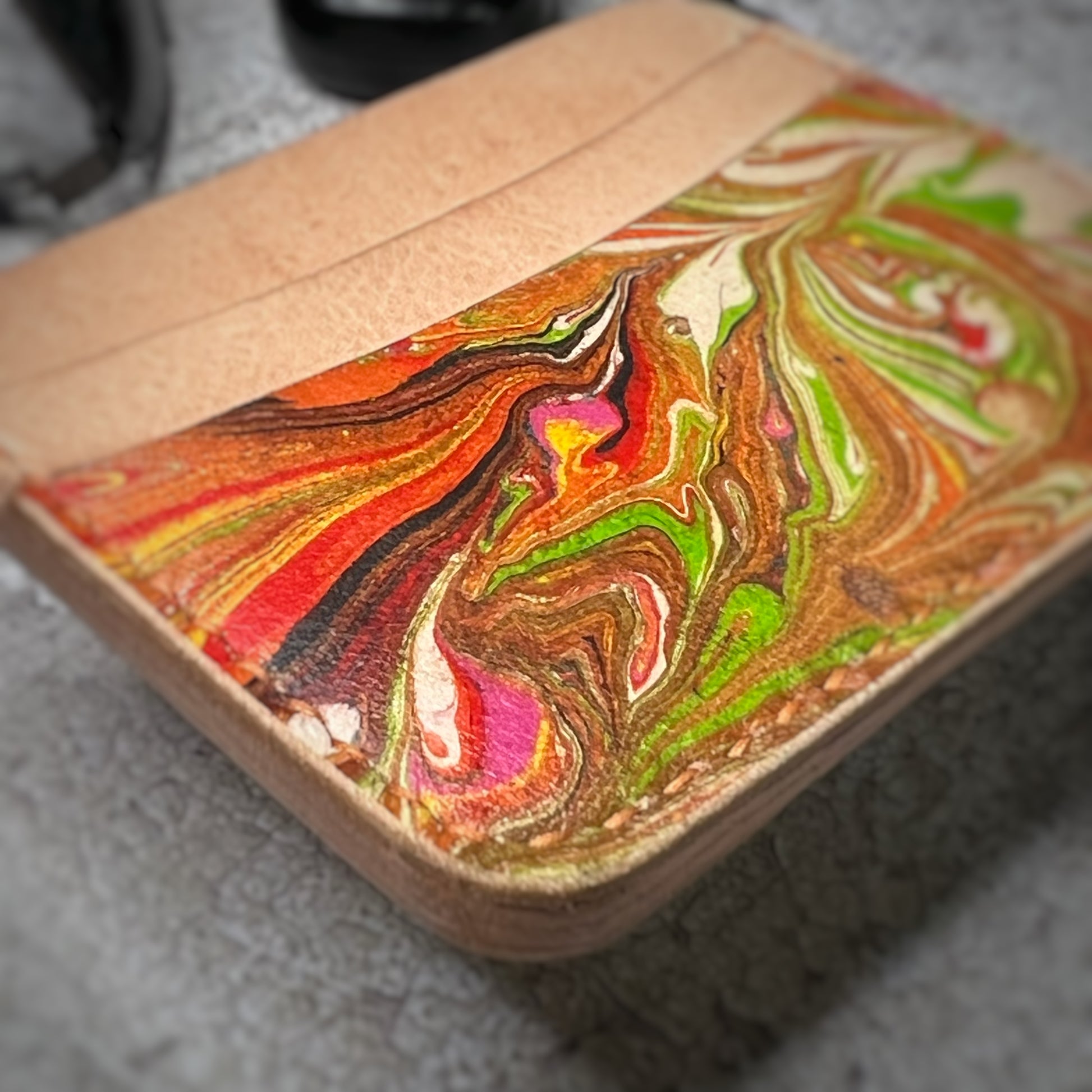 Close-up view of a leather cardholder with a natural tan upper section and a colorful, swirling pattern in green, red, and brown on the lower section. The cardholder is placed on a gray textured surface, showcasing the unique pattern.