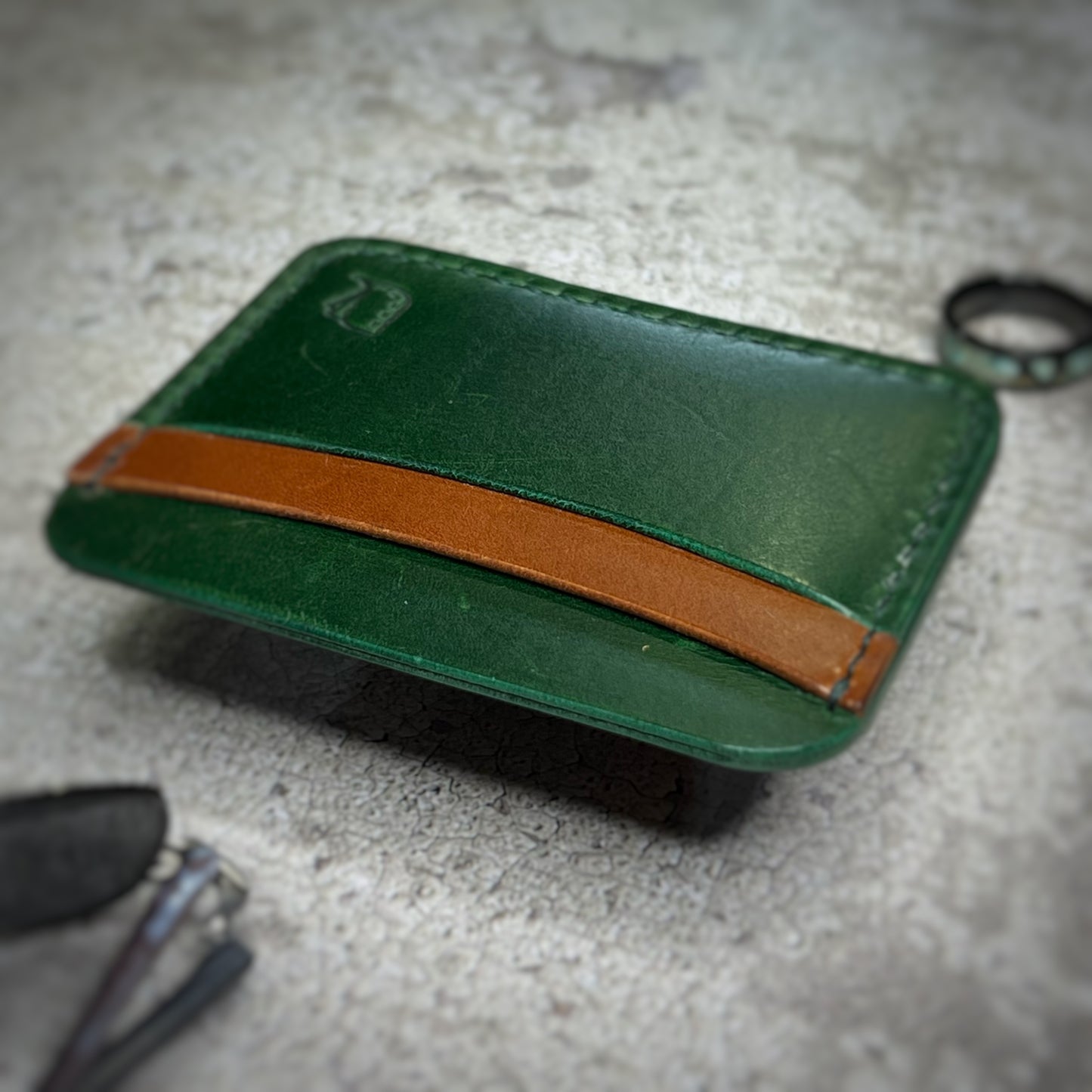 Close-up view of a leather cardholder with a green body and a brown strip across the top, showcasing the stitching and texture of the leather. The cardholder is placed on a gray textured surface.