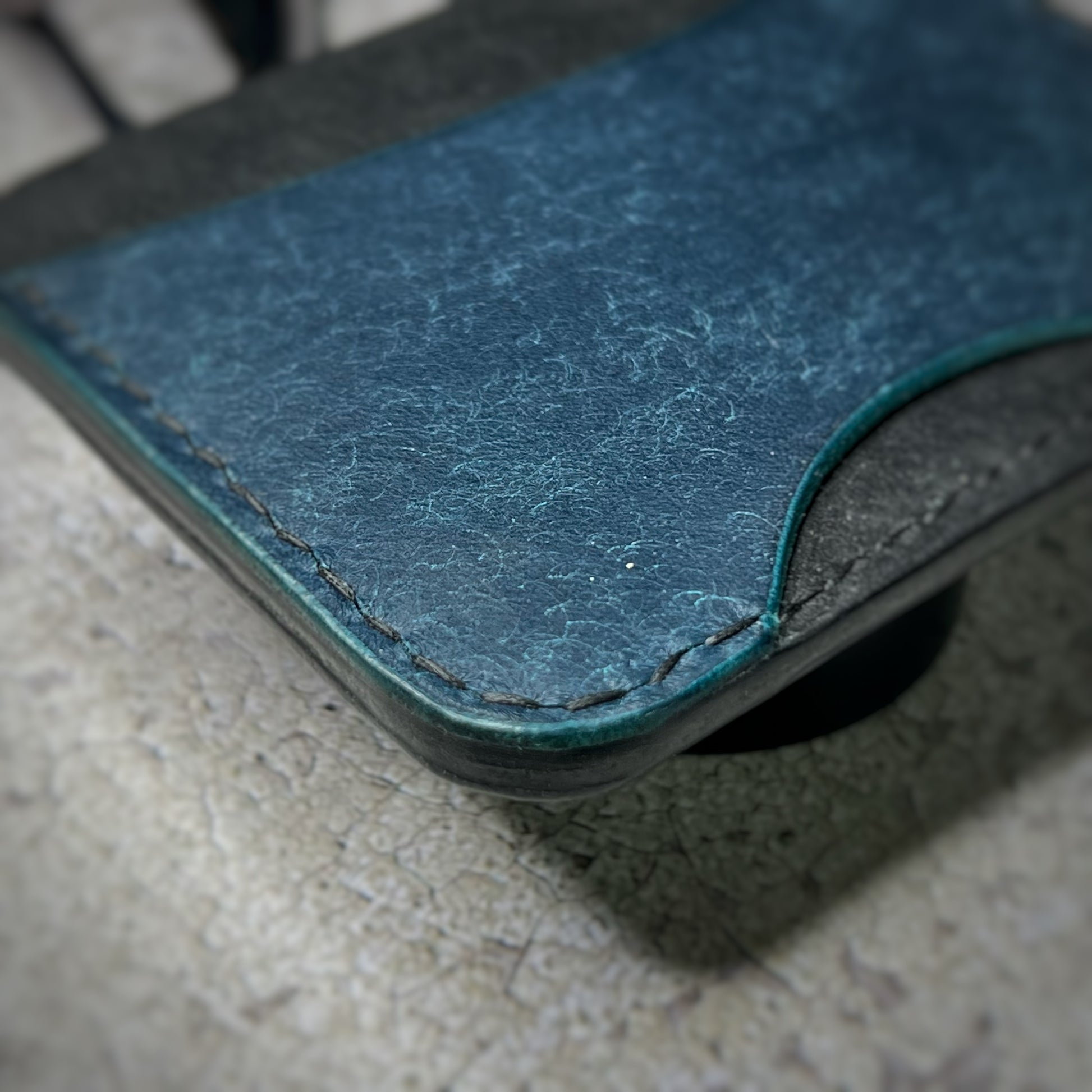 Close-up view of a leather cardholder with a black body and a blue section in the center, showcasing the stitching and texture of the leather. The cardholder is placed on a gray textured surface.