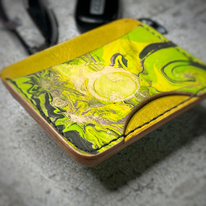 Close-up view of a leather cardholder with a yellow lower section, a black strip, and a vibrant green, swirling pattern on the upper section. The cardholder is placed on a gray textured surface, showcasing the unique pattern.