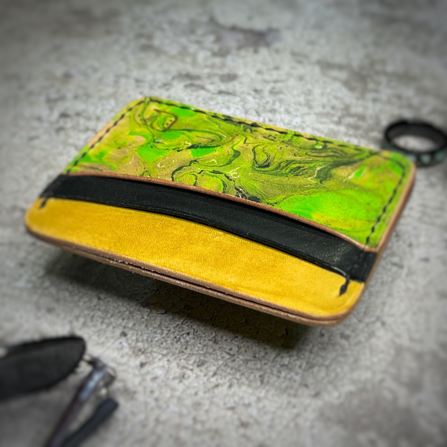 A leather cardholder with a yellow lower section, a black strip, and a vibrant green, swirling pattern on the upper section. The cardholder is placed on a gray textured surface.