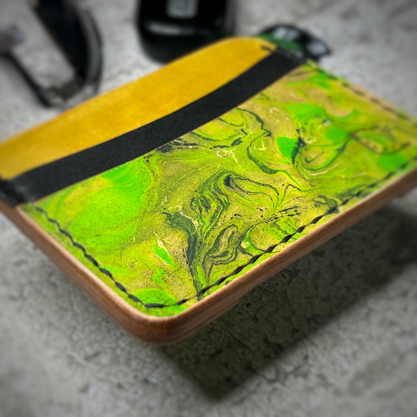 Close-up view of a leather cardholder with a yellow lower section, a black strip, and a vibrant green, swirling pattern on the upper section. The cardholder is placed on a gray textured surface.