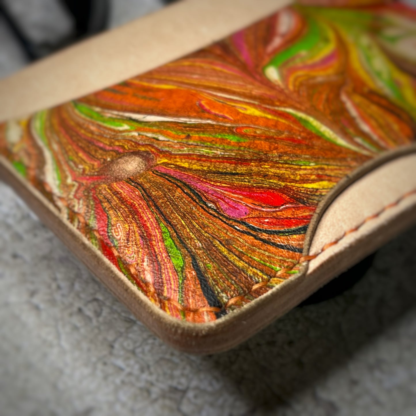 Close-up view of a leather cardholder with a natural tan upper section and a colorful, swirling pattern in green, red, and brown on the lower section. The cardholder is placed on a gray textured surface, highlighting the stitching details.