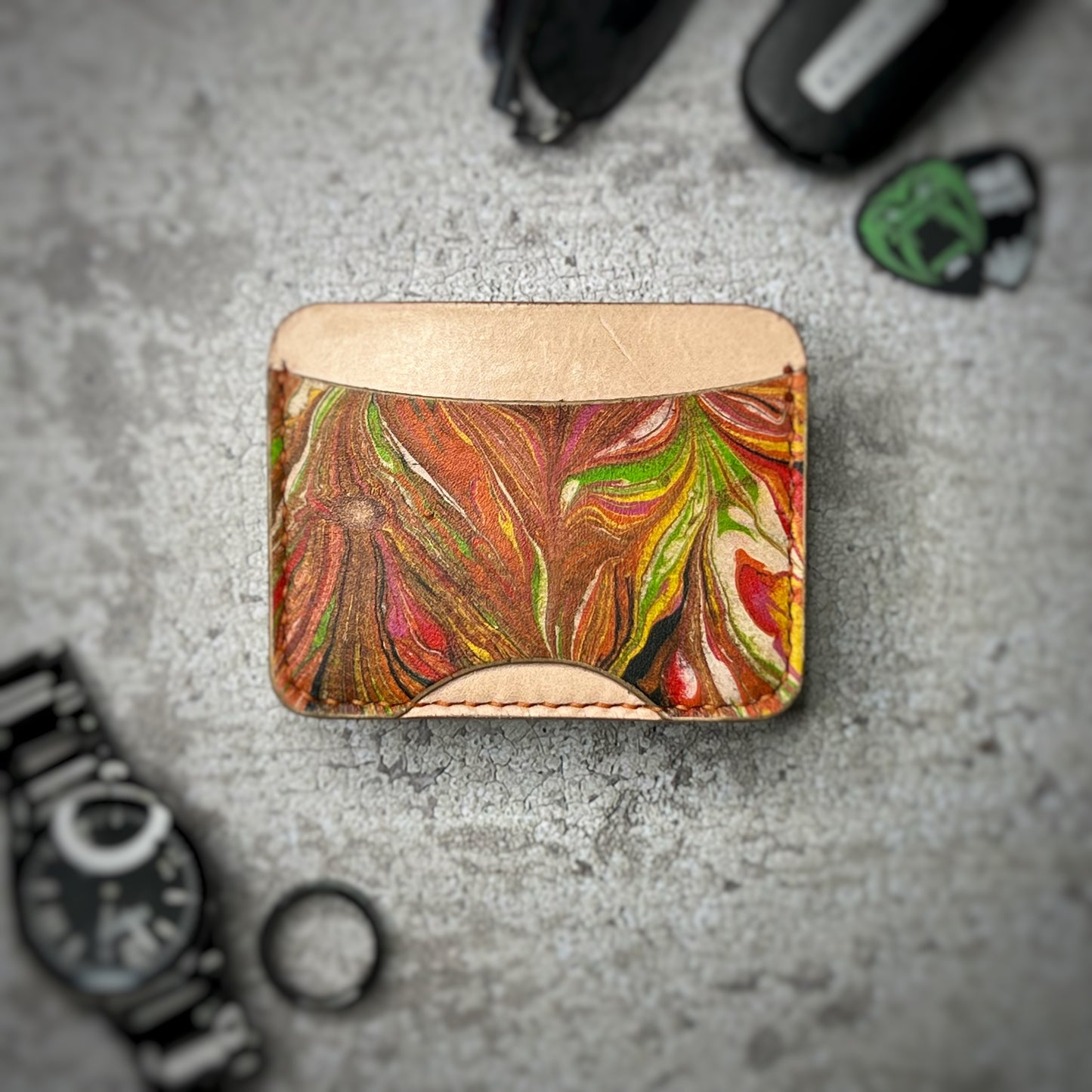 A leather cardholder with a natural tan upper section and a colorful, swirling pattern in green, red, and brown on the lower section. The cardholder is placed on a gray textured surface, surrounded by a black watch, a black ring, and some keys and other small objects in the background.