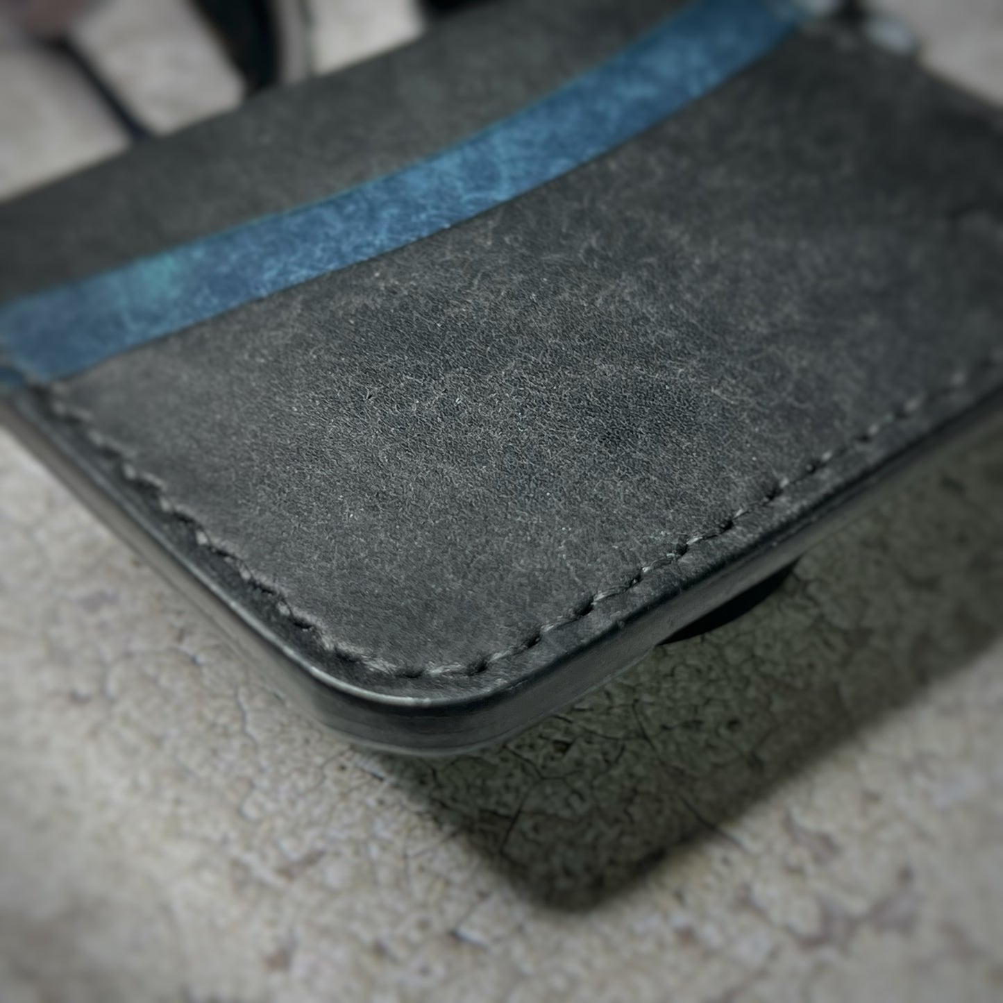 Close-up view of a leather cardholder with a black body and a blue strip across the top, showcasing the stitching details. The cardholder is placed on a gray textured surface.