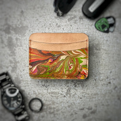 A leather cardholder with a natural tan upper section and a colorful, swirling pattern in green, red, and brown on the lower section. The cardholder is placed on a gray textured surface, surrounded by a black watch, a black ring, and some keys and other small objects in the background.