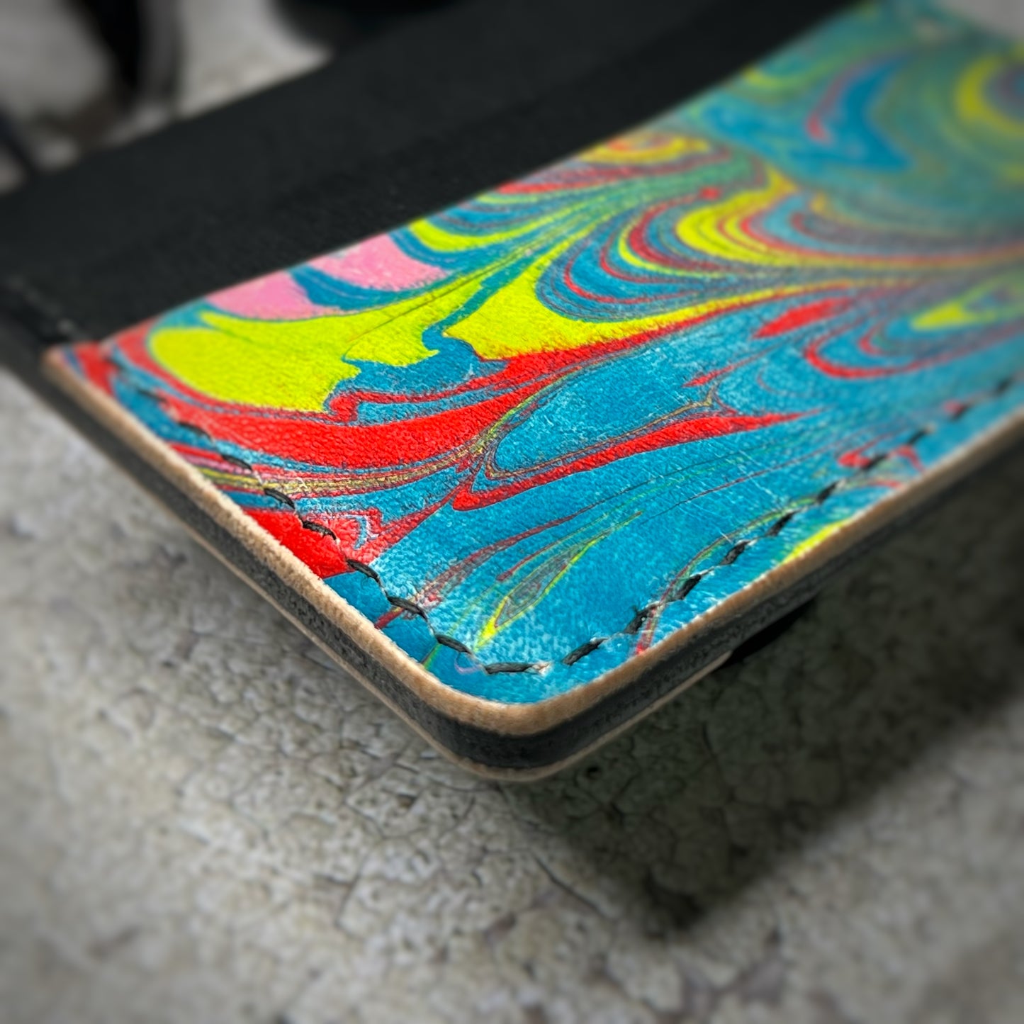 Close-up view of a colorful leather cardholder with a black upper section and a vibrant, swirling pattern in yellow, blue, red, and green on the lower section. The cardholder is placed on a gray textured surface, showcasing the corner details.