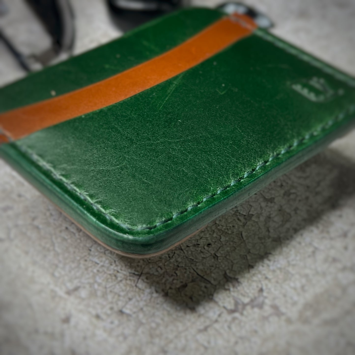 A leather cardholder with a green upper section and a brown lower section, featuring a minimalist design. The cardholder is placed on a gray textured surface, surrounded by a black watch, a black ring, and some keys and other small objects in the background.