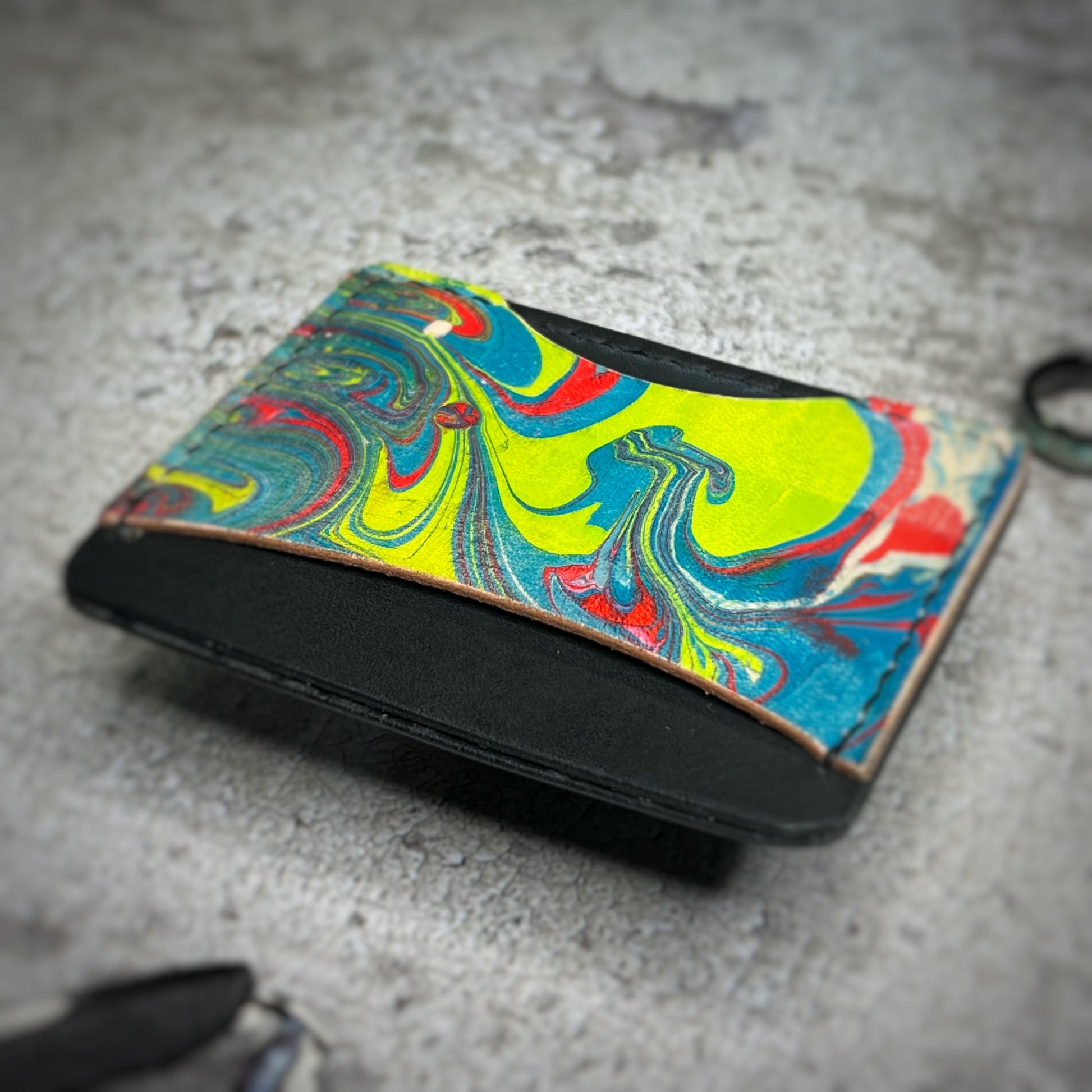 A colorful cardholder made of leather with a black upper section and a vibrant, swirling pattern in yellow, blue, red, and green on the lower section. The cardholder is placed on a gray textured surface, showcasing a slightly angled view.