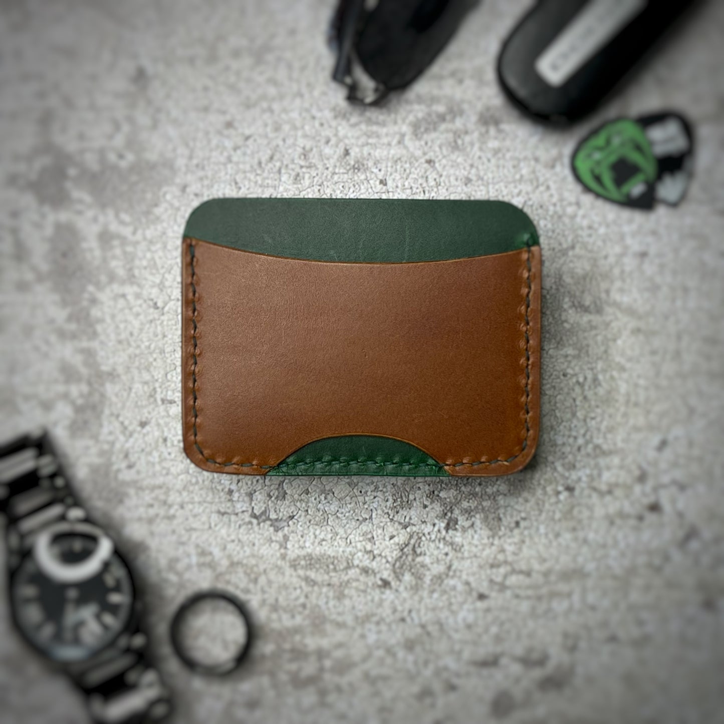 A leather cardholder with a green upper section and a brown lower section. The cardholder is placed on a gray textured surface, surrounded by a black watch, a black ring, and some keys and other small objects in the background.