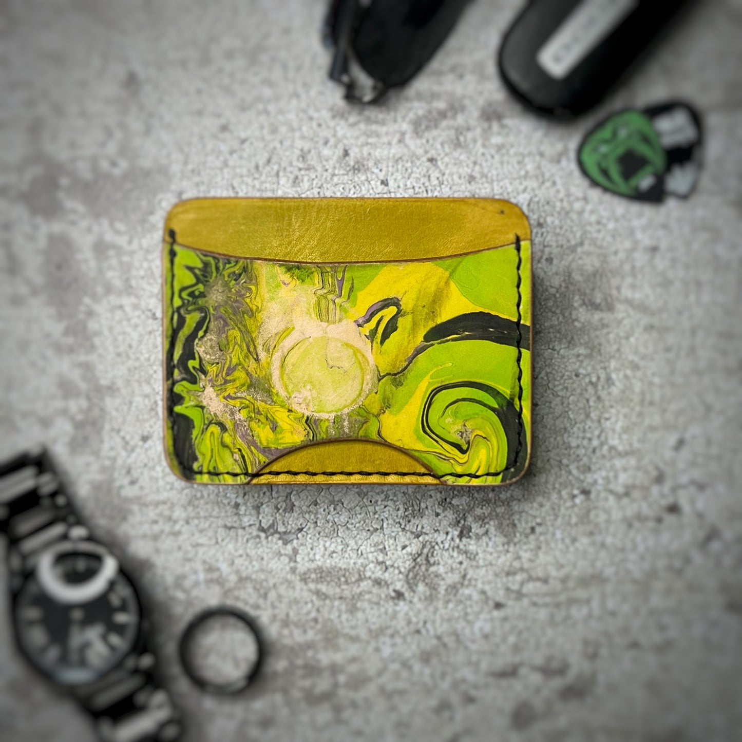 A leather cardholder with a yellow upper section, a black strip, and a vibrant green, swirling pattern on the lower section. The cardholder is placed on a gray textured surface, surrounded by a black watch, a black ring, and some keys and other small objects in the background.