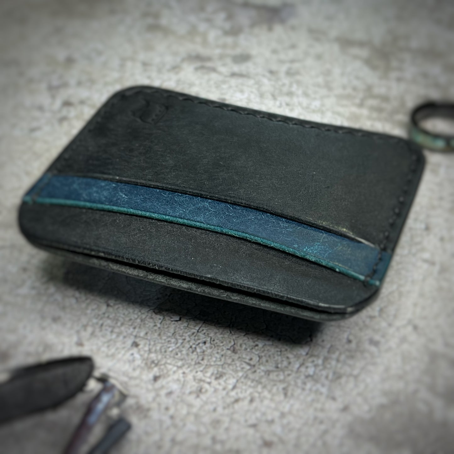 A leather cardholder with a black body and a blue strip across the top. The cardholder is placed on a gray textured surface with some keys and other small objects in the background.