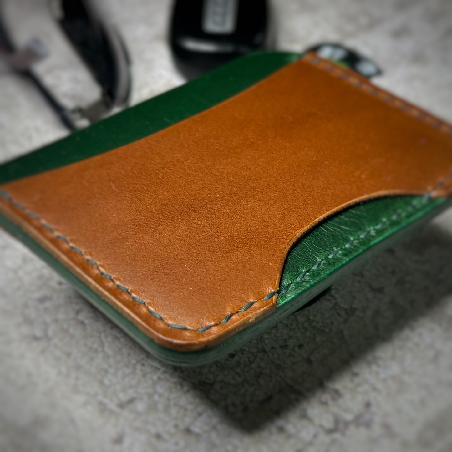 Close-up view of a leather cardholder with a green body and a brown strip across the top, showcasing the stitching details. The cardholder is placed on a gray textured surface.