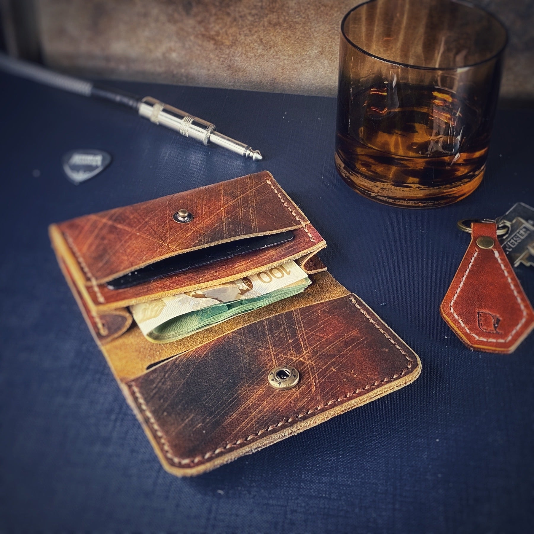 A locust card holder made from distressed leather dyed English tan with tan stitching. Around the wallet are a leather keyfob, a glass with some liquid in it, the end of a guitar cord and a Metallica guitar pick.