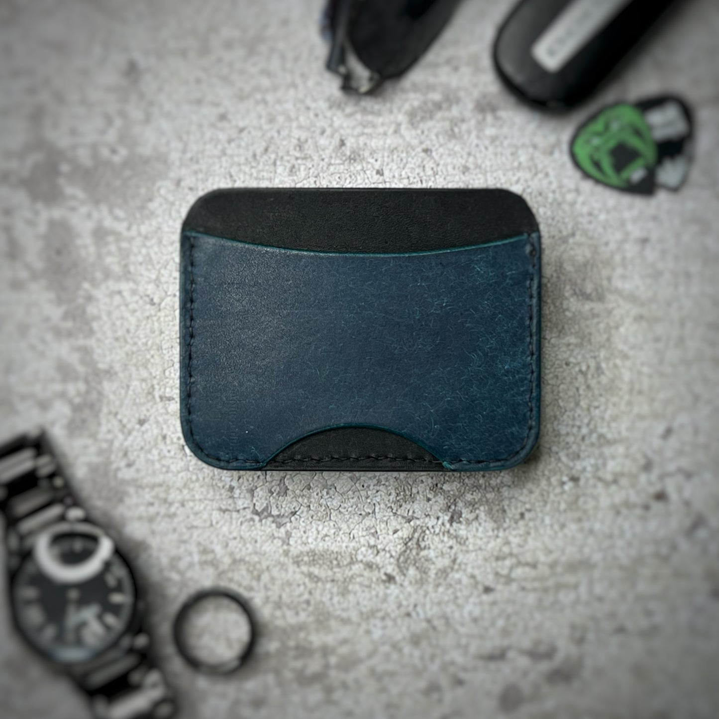 A leather cardholder with a black upper section and a blue lower section, showcasing a simple and elegant design. The cardholder is placed on a gray textured surface, surrounded by a black watch, a black ring, and some keys and other small objects in the background.