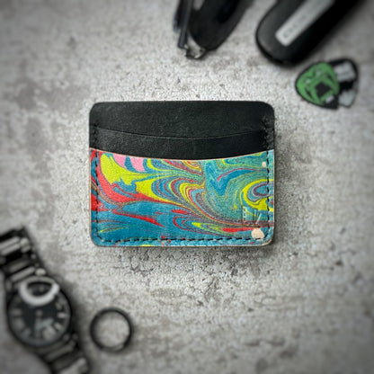 A colorful cardholder made of leather with a black upper section and a vibrant, swirling pattern in yellow, red, blue, and green on the lower section. The cardholder is placed on a gray textured surface, surrounded by a black watch, a black ring, and some keys and other small objects in the background.