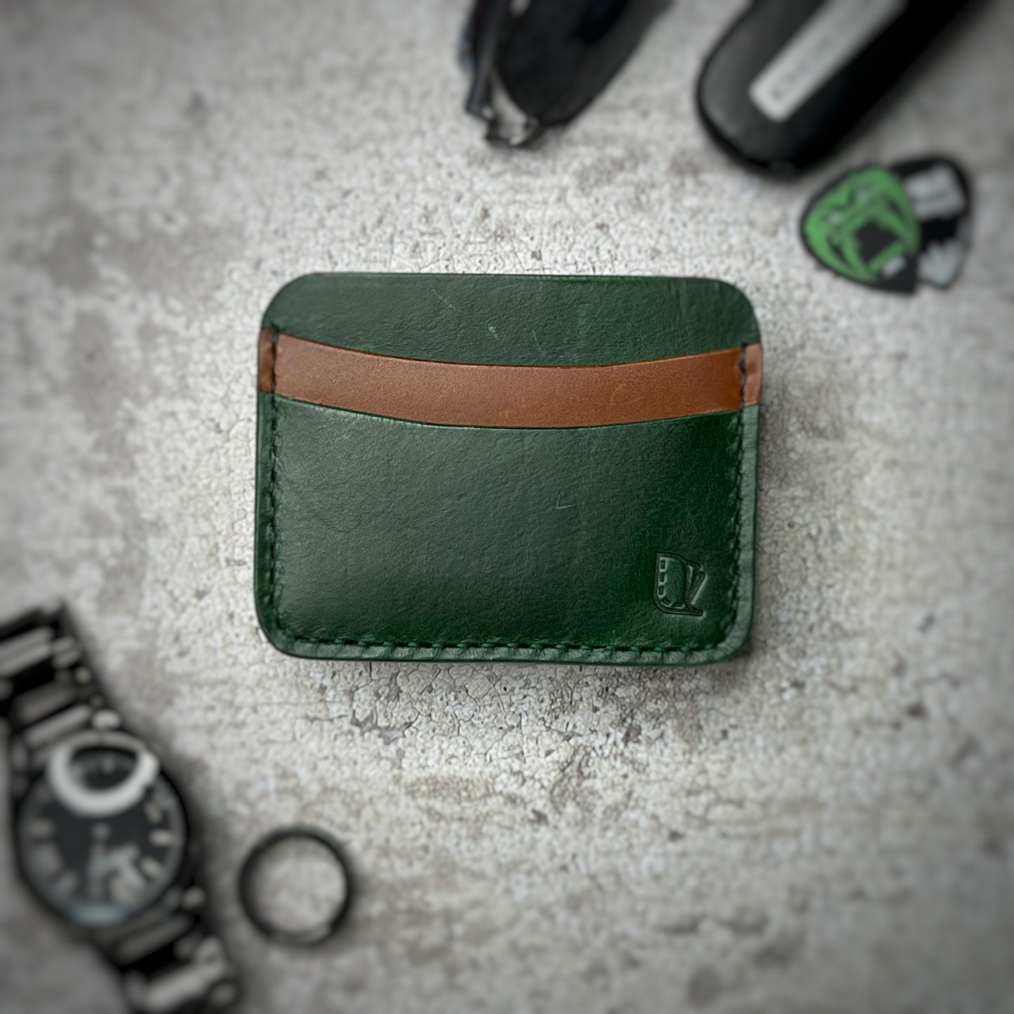 A leather cardholder with a dark green body and a brown strip across the top. The cardholder is placed on a gray textured surface, surrounded by a black watch, a black ring, and some keys and other small objects in the background.