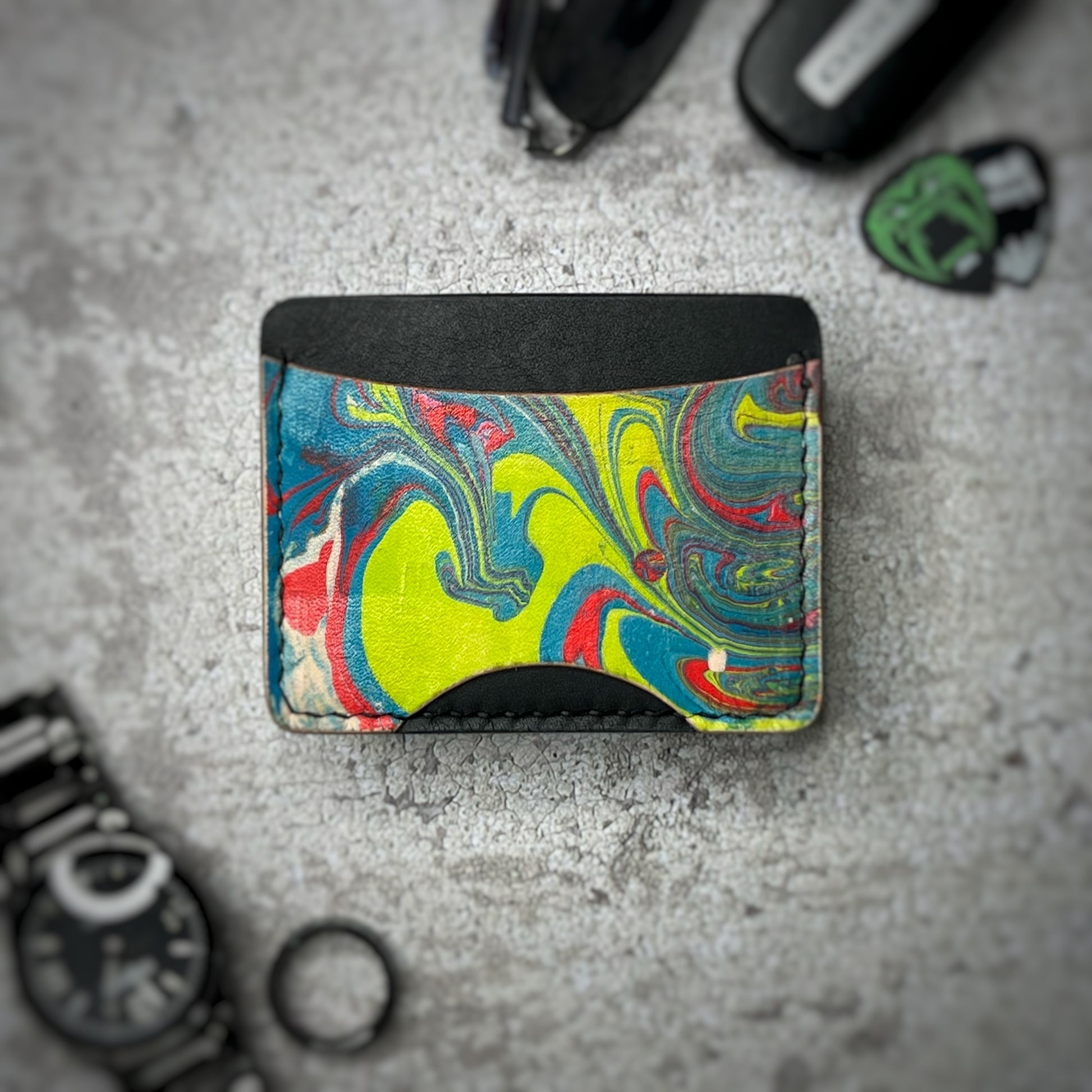 A colorful cardholder made of leather with a black upper section and a vibrant, swirling pattern in yellow, blue, red, and green on the lower section. The cardholder is placed on a gray textured surface, surrounded by a black watch, a black ring, and some keys and other small objects in the background.