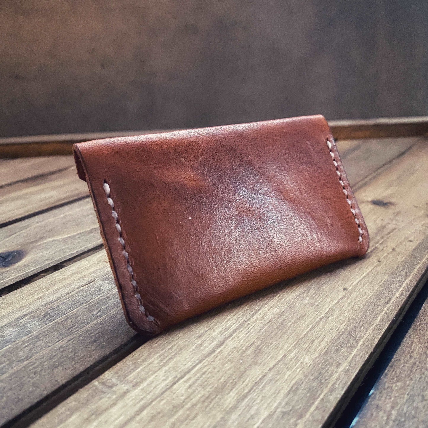 The back of a flap top leather card holder in saddle tan with white stitching sitting on a dark wood platform.