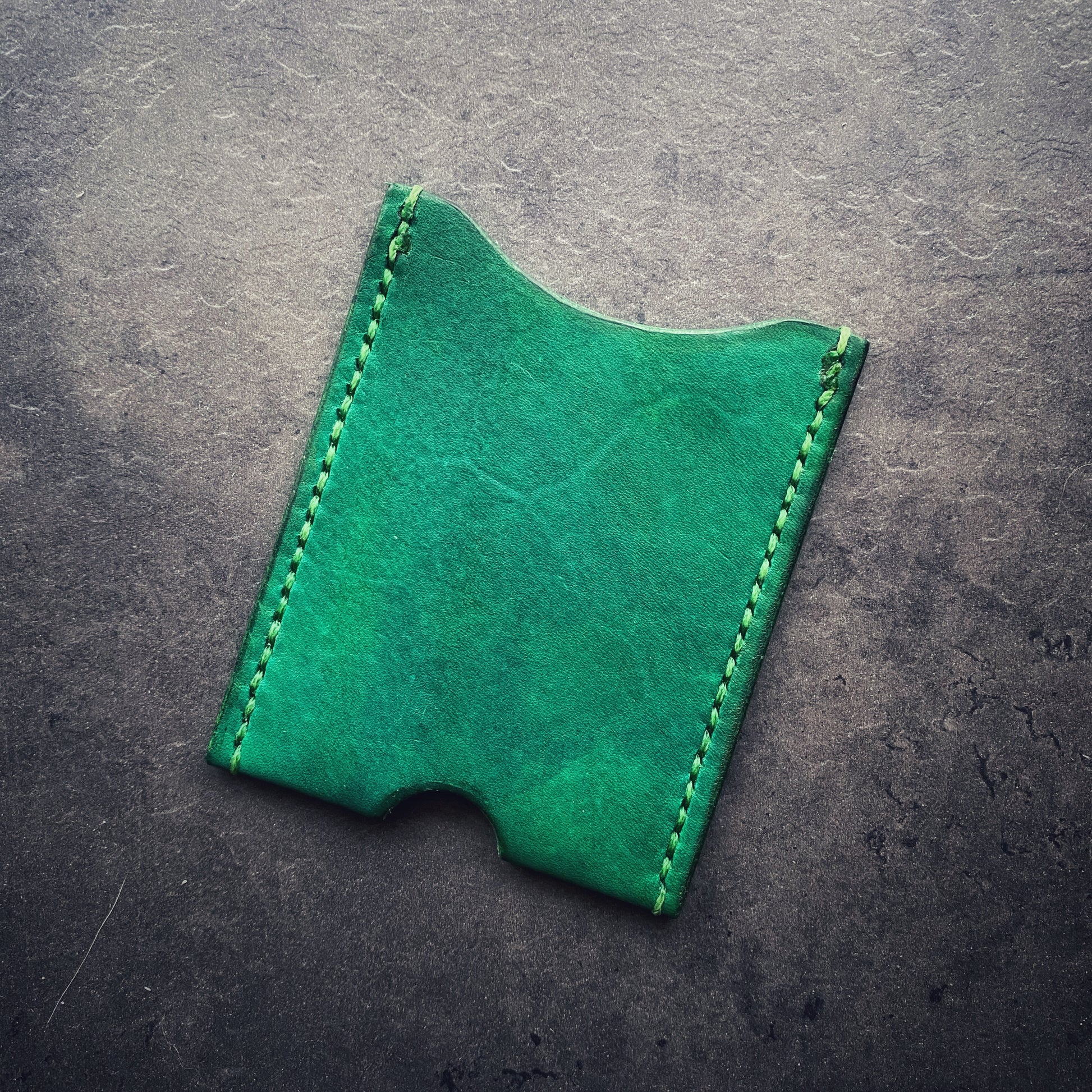 The elm leather cardholder in Kelly green with green stitching sitting on a concrete background. The cardholder is rectangular in shape with a semicircular thumb notch in the middle of the bottom seam. 