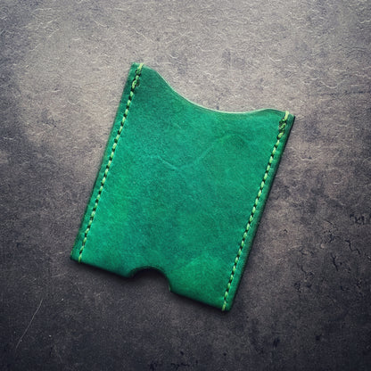 The elm leather cardholder in Kelly green with green stitching sitting on a concrete background. The cardholder is rectangular in shape with a semicircular thumb notch in the middle of the bottom seam. 